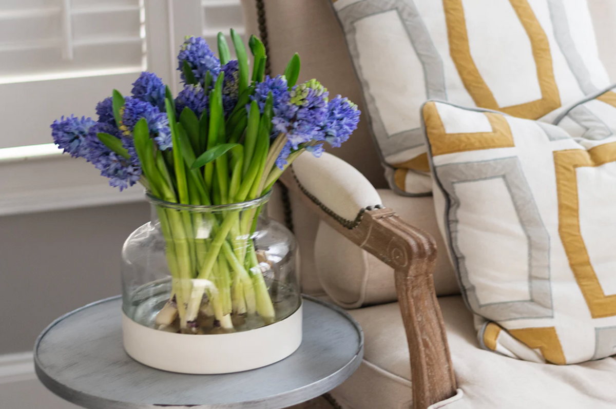 Clear vase with blue flowers on a small round table next to a striped armchair.